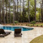 Kidney-shaped pool on a stone patio with two brown wicker lounge chairs; tall trees and a fence in the background with greenery beyond.