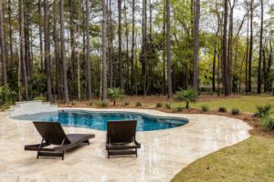 Kidney-shaped pool on a stone patio with two brown wicker lounge chairs; tall trees and a fence in the background with greenery beyond.
